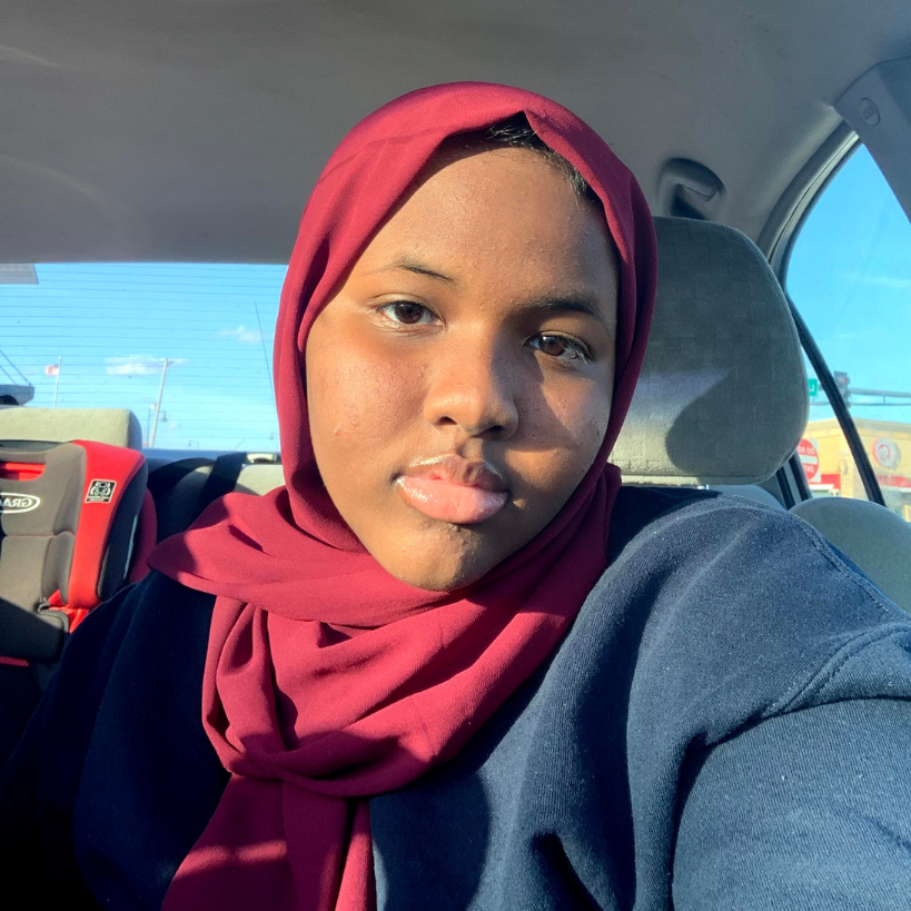Female with dark top and red hijab looks into the camera. They are in a car. 