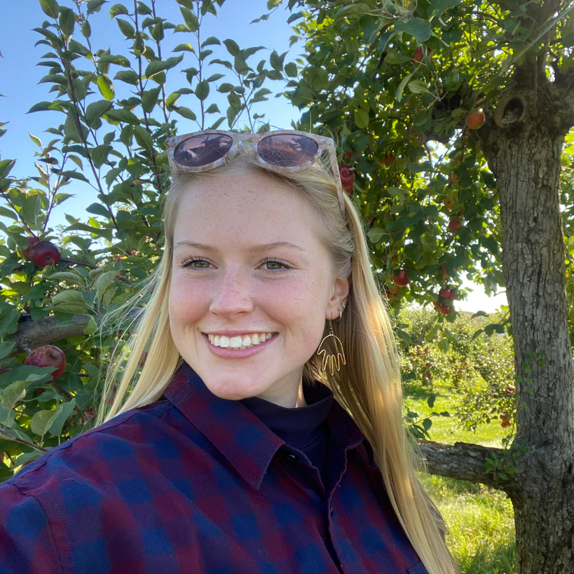 female with light hair standing in front of a tree. Smiles at camera, has sunglasses on top of head. Wearing a blue and purple checked shirt.