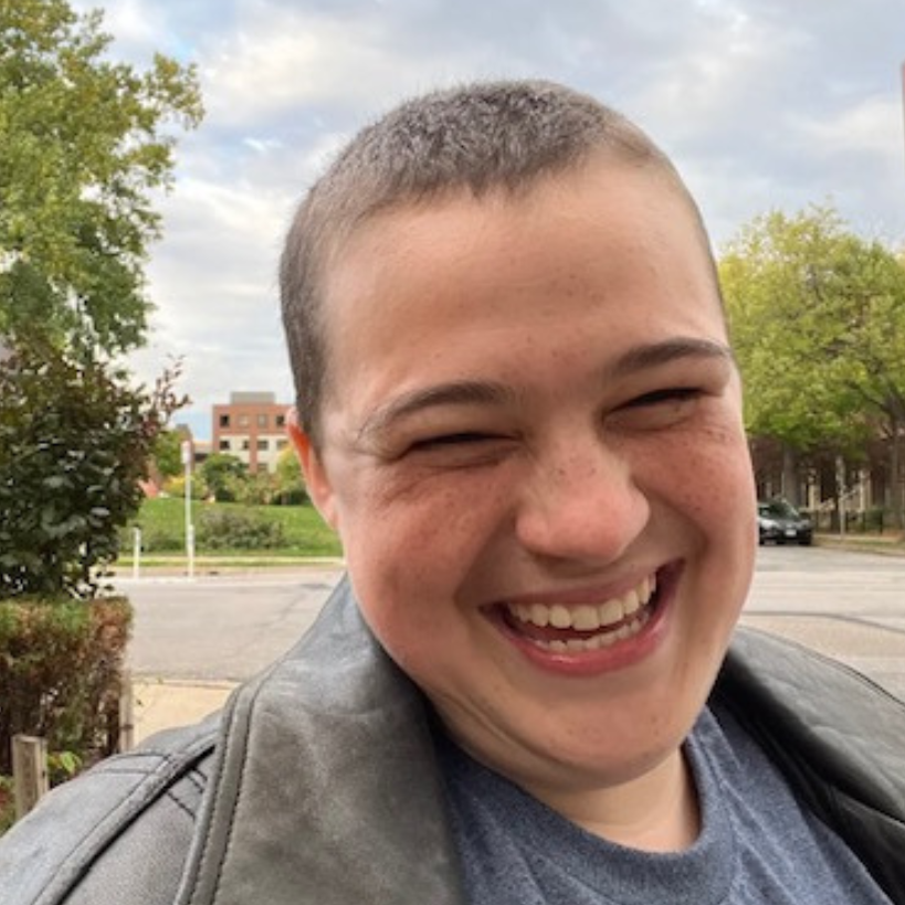  person with shaved head smiles at the camera. they appear to be outdoors with trees and a street in the background.