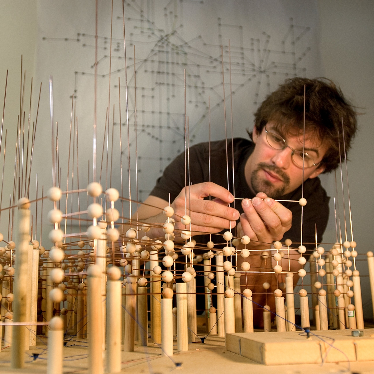 A student with brown hair, beard, and glasses looks intently at a complex wood and wire structure. The structure is made of vertical copper wires, small wooden balls, and short wooden dowels. 