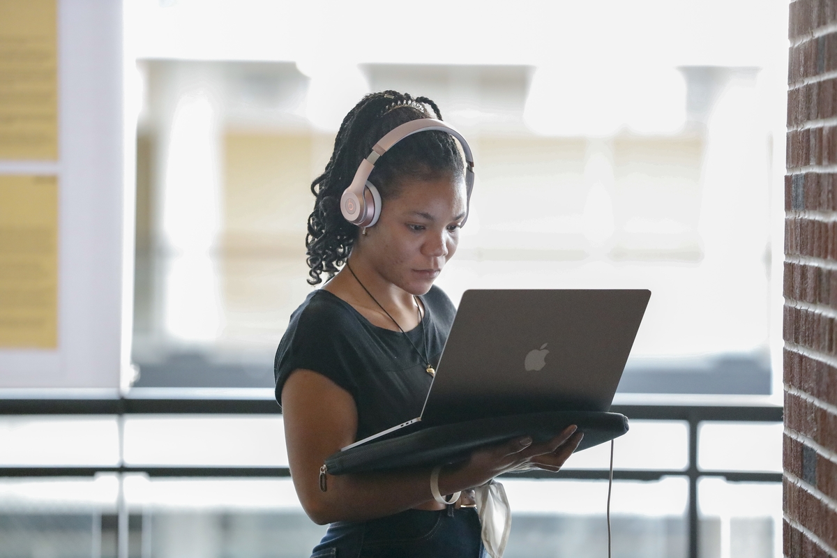 person with dark skin holds a laptop and looks at it while wearing headphones