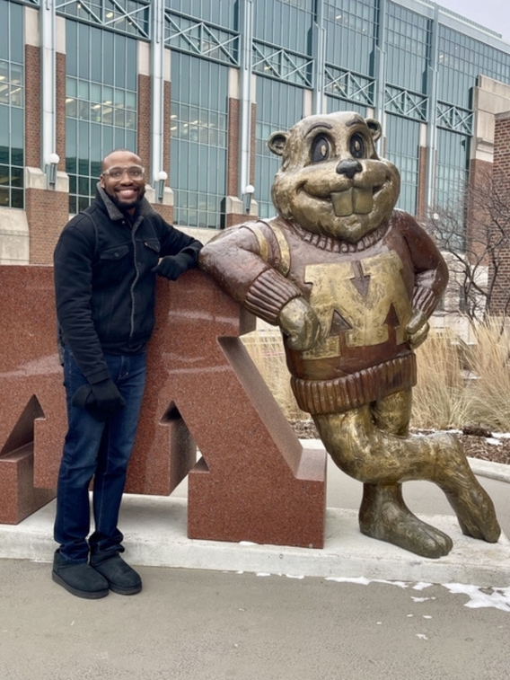 person with dark skin and glasses smiles at the camera, posing next to Goldy statue.